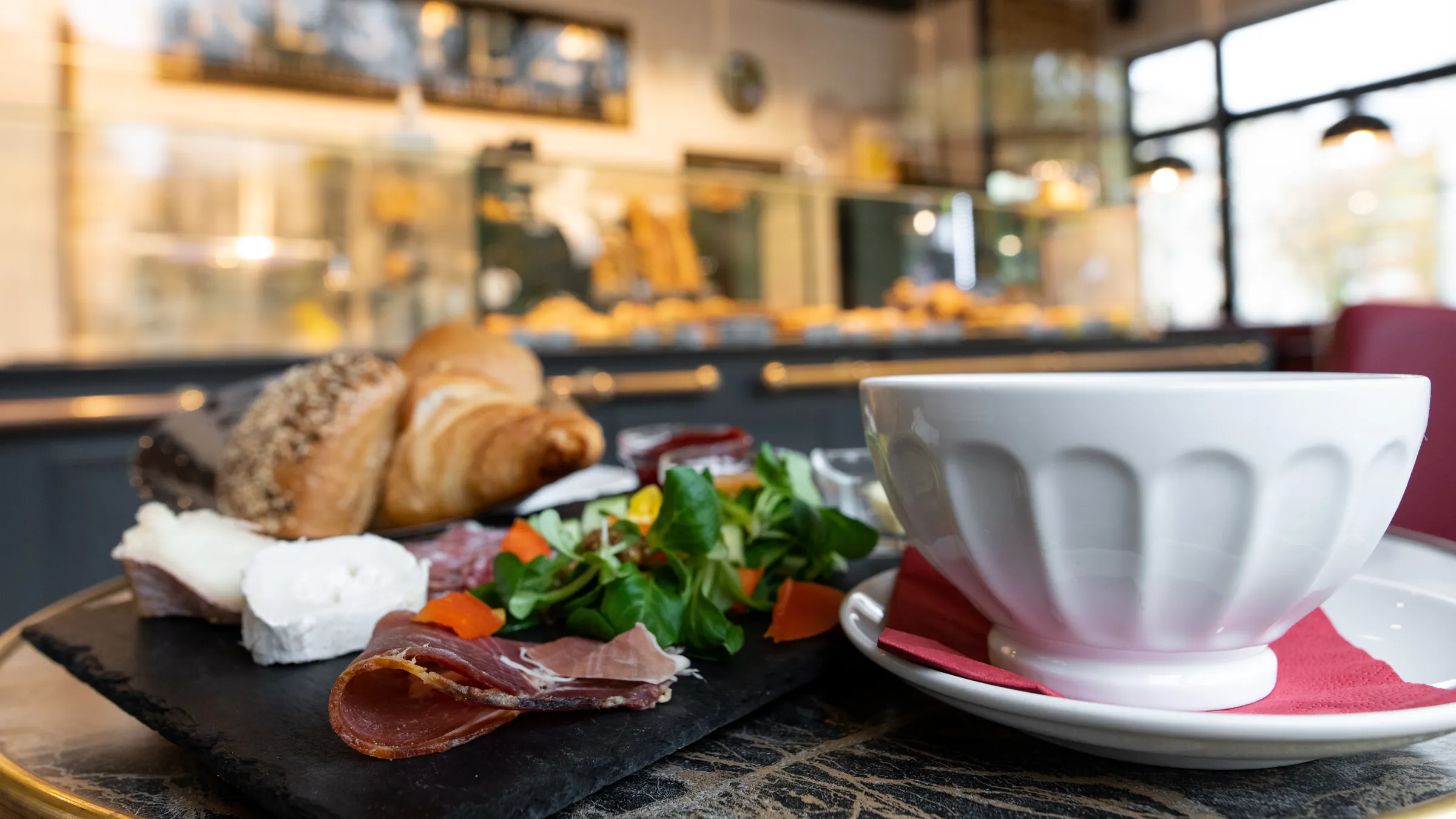Schieferplatte mit Rohschinken, Ziegenkäse, Salat und Brötchen auf dem Bistrotisch, daneben eine Kaffeeschale, dahinter die Theke